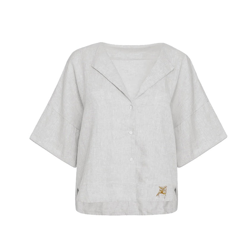 Image of a white linen shirt with short sleeves and a wing-style collar. The shirt has embroidery on the bottom of each side. The purpose of this image is to highlight the shirt by NAOS because it's for sale at EVAMAIA Boutique in Studio City, CA and online.