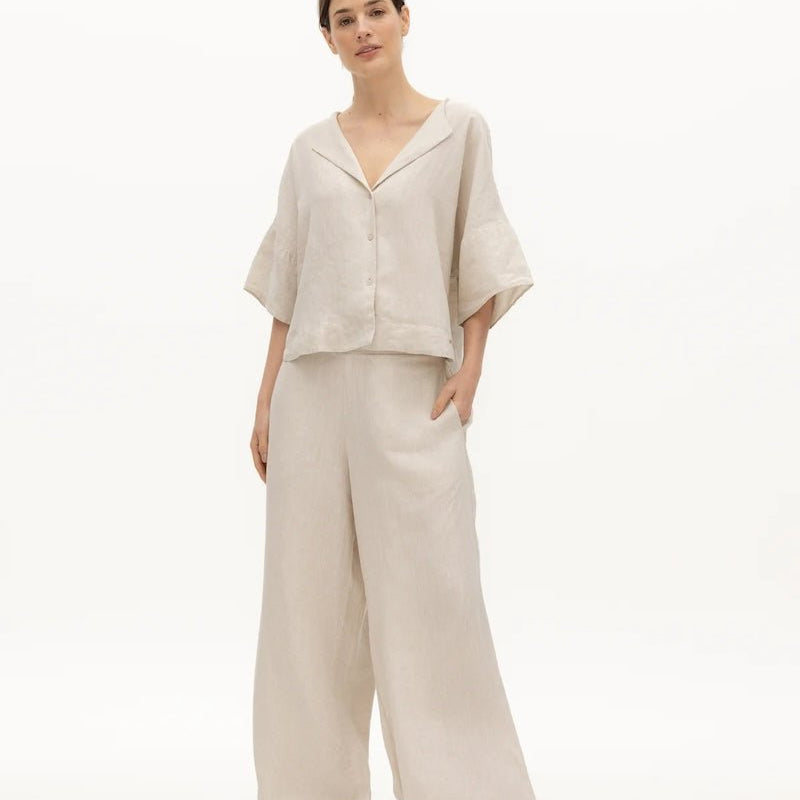 Image of a woman wearing a sand-colored linen top and a matching bottom. The top has a wing collar and the bottom is long and flowy. The purpose of the image is to highlight the clothing by NAOS because it's available for sale at EVAMAIA boutique in Studio City, CA.