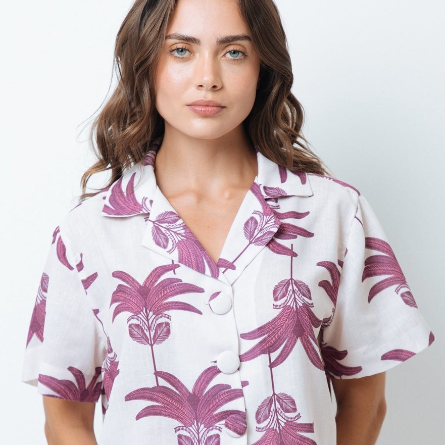 Woman wearing a shirt with pink palm trees. She's standing behind a white background. The purpose of this image is to highlight the shirt by Mar de Lua.