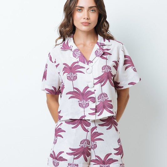 Woman wearing a shirt with pink palm trees and a matching bottom. She's standing behind a white background. The purpose of this image is to highlight the clothing by Mar de Lua.