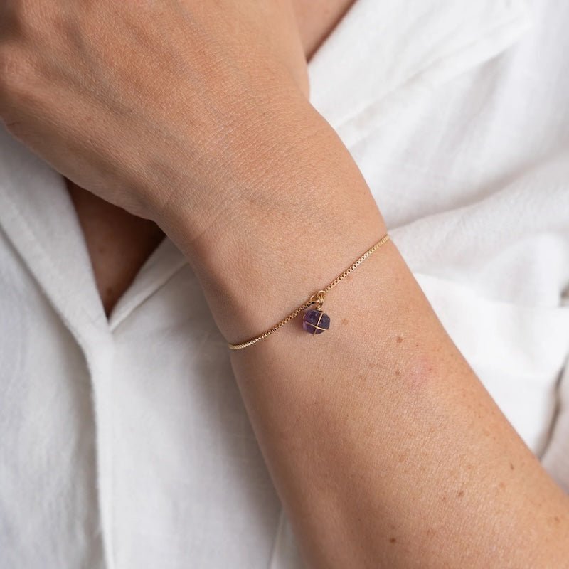 Close up image of a woman wearing a gold chain bracelet with an amethyst stone on it. The purpose of the image is to highlight the bracelet because it is on sale at EVAMAIA boutique in Studio City, CA.