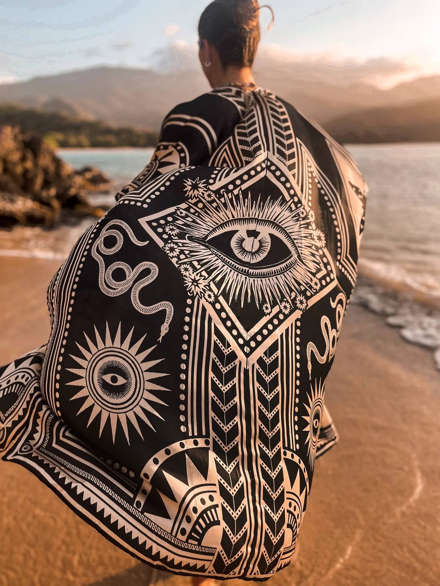 The designer of PLISSE, Paloma wearing the nazar kimono and wrap skirt. She is posing in the sand at a beach during golden hour. This is the back of this flowy kimono.