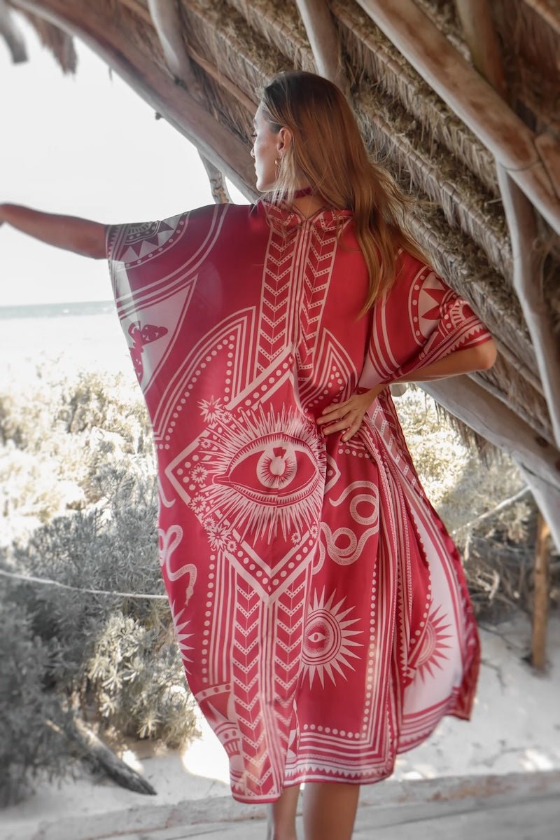 Image of a woman wearing a light red kimono with the image of an eye, snakes, and the sun. The purpose of this image is to highlight the kimono, which is for sale at EVAMAIA boutique in studio city, CA.