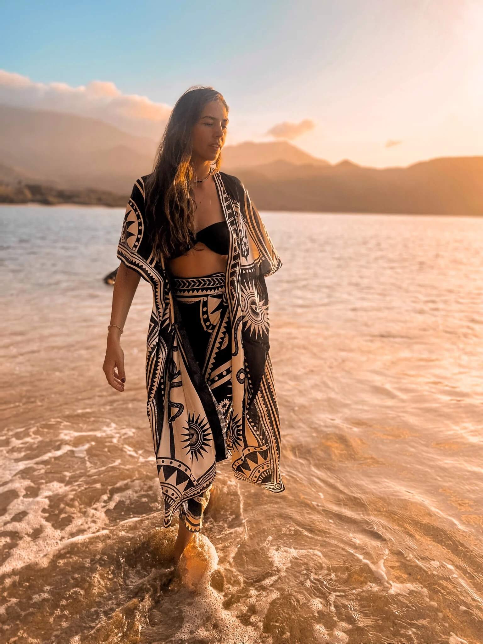 The designer of PLISSE, Paloma wearing the nazar kimono and wrap skirt. She is posing in the water at a beach during golden hour. This is the front of this flowy kimono.