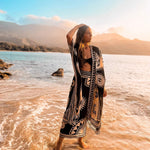 The designer of PLISSE, Paloma wearing the nazar kimono and wrap skirt. She is posing with her arms on her head in the sand at a beach during golden hour. This is the front of this flowy kimono.