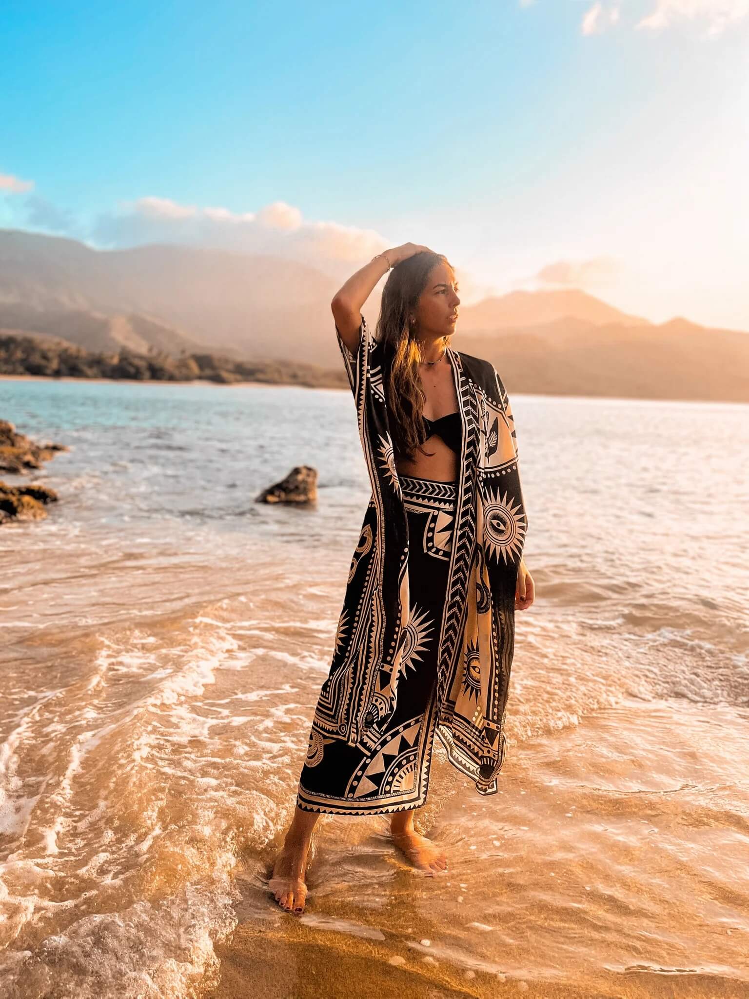 The designer of PLISSE, Paloma wearing the nazar kimono and wrap skirt. She is posing with her arms on her head in the sand at a beach during golden hour. This is the front of this flowy kimono.