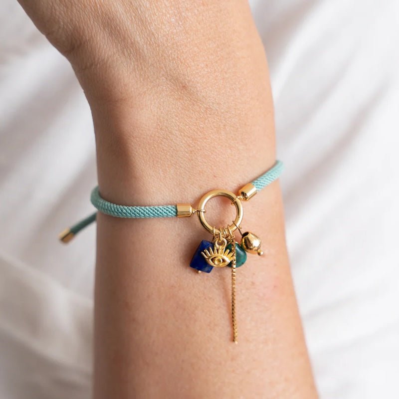 Close-up image of a woman wearing a thread bracelet with many charms including an eye and a green and blue stone. The purpose of this image is to promote the bracelet because it is for sale at EVAMAIA boutique in studio city, ca and online.