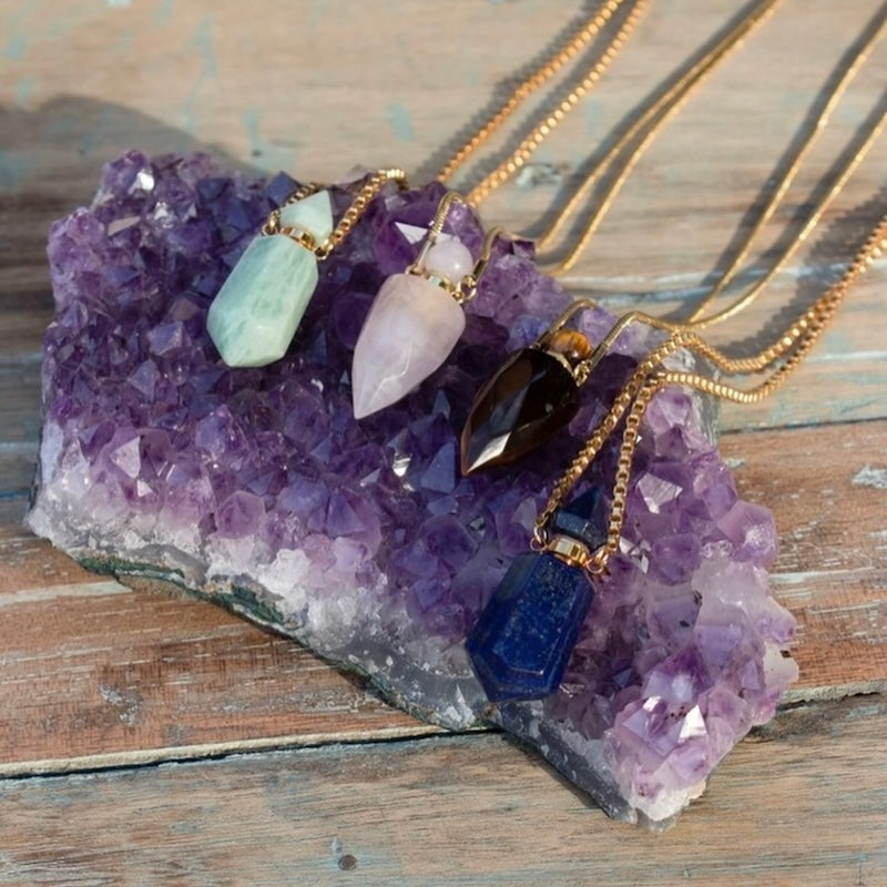 Image of perfume bottles made out of stone sitting on a raw amethyst stone. The purpose of this image is to highlight the perfume bottles, which can be worn as necklaces. They are made by Pajarolimon and sold at EVAMAIA boutique.