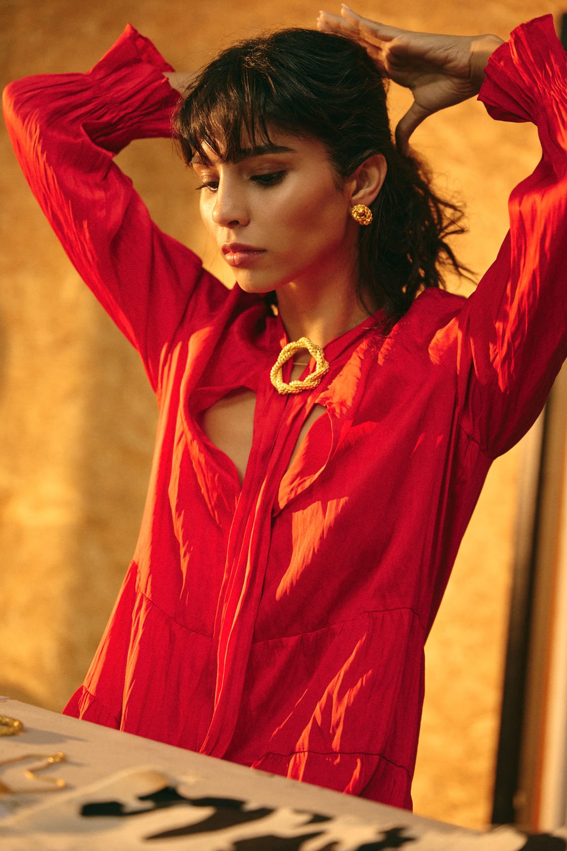 Image of a woman wearing gold earrings and a matching pin. She's also wearing a red dress and seems to be getting ready, putting her hair back. The purpose of this image is to highlight the earrings by Alma jewelry.