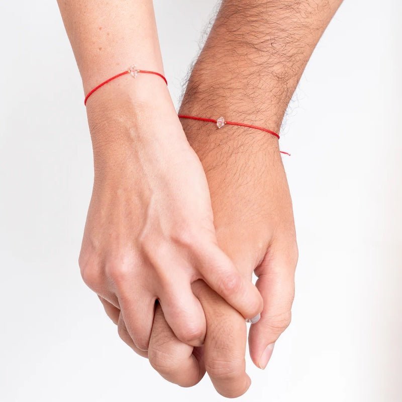 Close up image of a man and a woman wearing a red threaded bracelet with a clear stone on it. A Herkimer diamond. The purpose of this image is to highlight the bracelet because it is for sale at EVAMAIA in Studio City, CA and online.