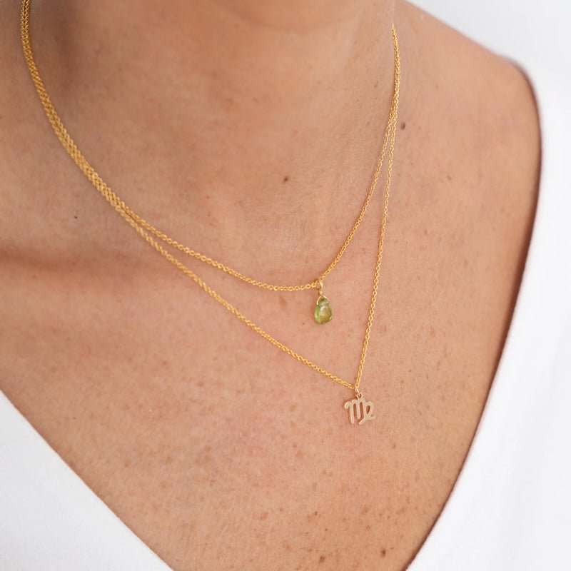 Close up image of a woman's neckline. She's wearing two necklaces. One has a charm of a Virgo zodiac sign and the other has a peridot stone. The purpose of this image is to highlight the necklaces, which are made in Colombia and sold in Studio City, at EVAMAIA boutique.