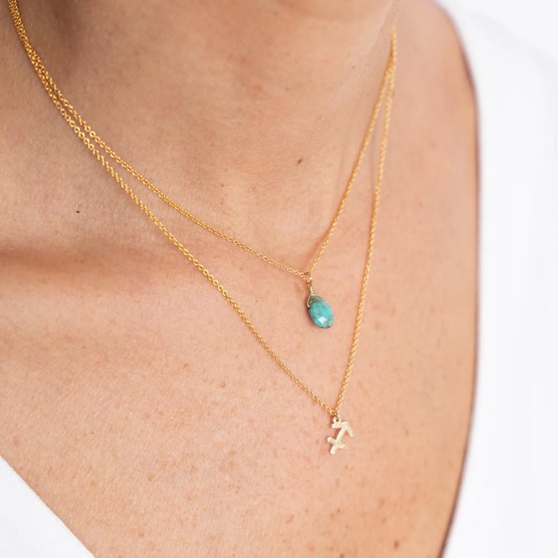 Close up image of a woman's neckline. She's wearing two necklaces. One has a charm of a Scorpio zodiac sign and the other has a Turquoise stone. The purpose of this image is to highlight the necklaces, which are made in Colombia and sold in Studio City, at EVAMAIA boutique.