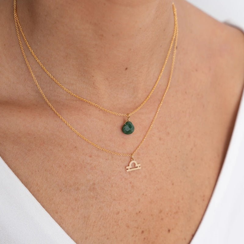 Close up image of a woman's neckline. She's wearing two necklaces. One has a charm of a Libra zodiac sign and the other has a green quartz. The purpose of this image is to highlight the necklaces, which are made in Colombia and sold in Studio City, at EVAMAIA boutique.